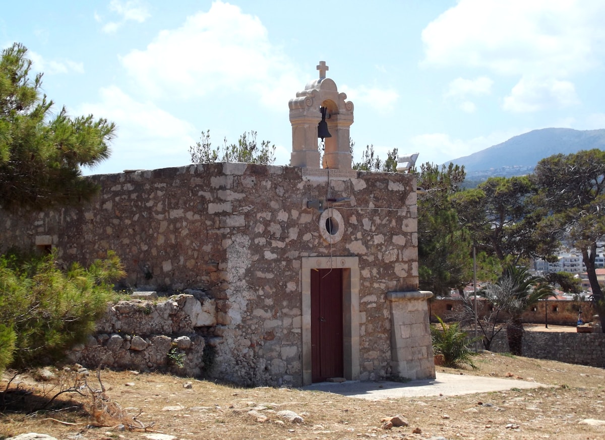 Small stone chapel with bell tower surrounded by pine trees and mountains in the Cretan countryside