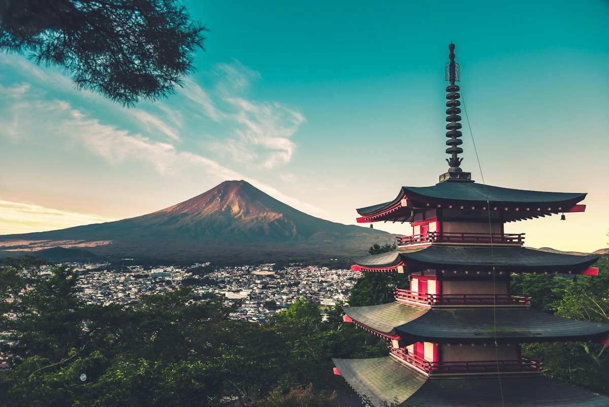 Red five storey Chureito Pagoda on a forested hillside with the snow capped cone of Mount Fuji behind it