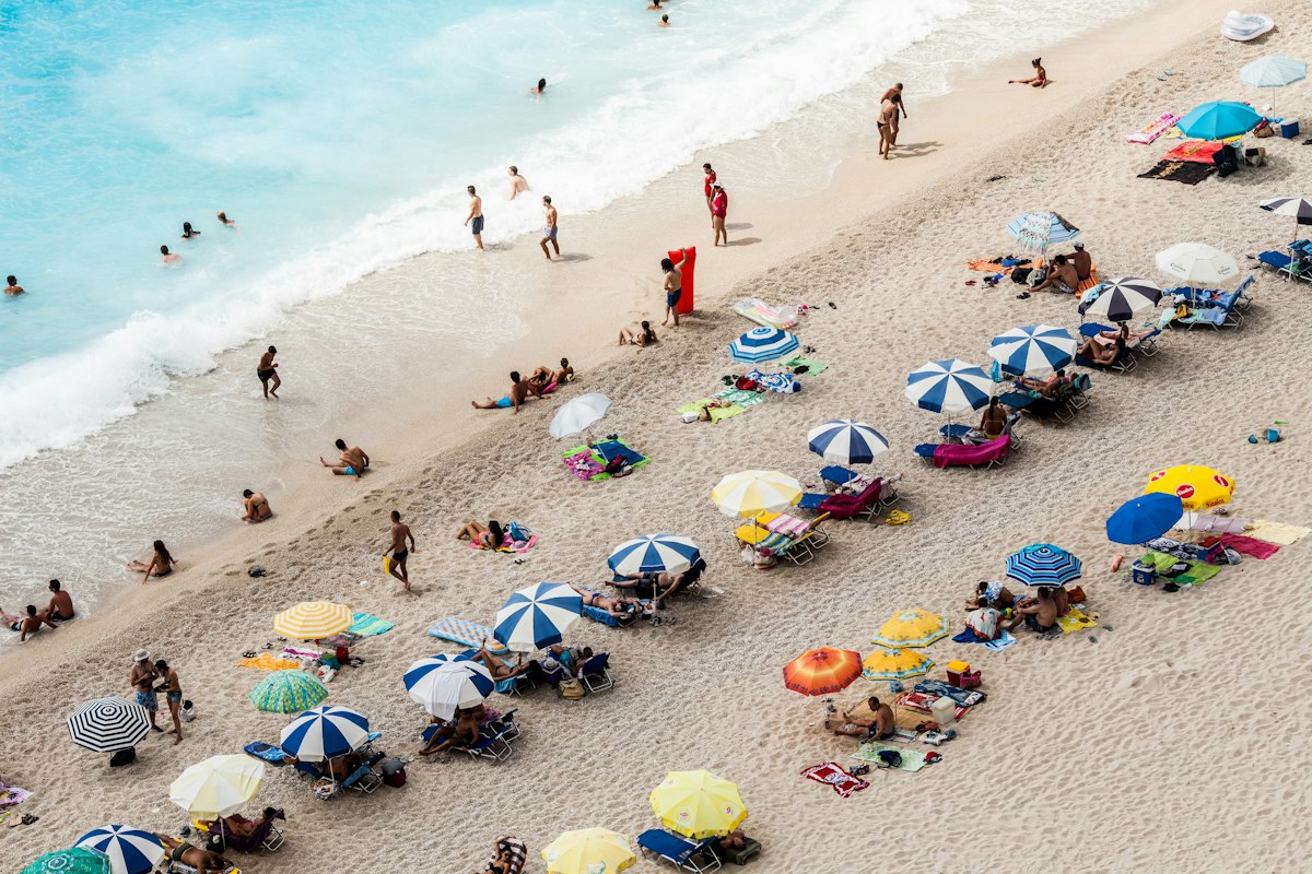 Aerial view of a crowded summer beach packed with colorful umbrellas and swimmers in shallow blue water