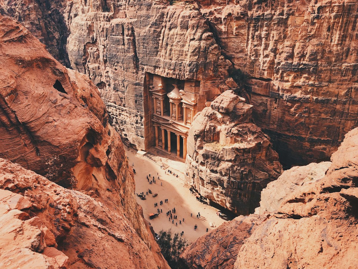 Aerial view of the Treasury at Petra carved into red sandstone cliffs with visitors standing in the canyon below
