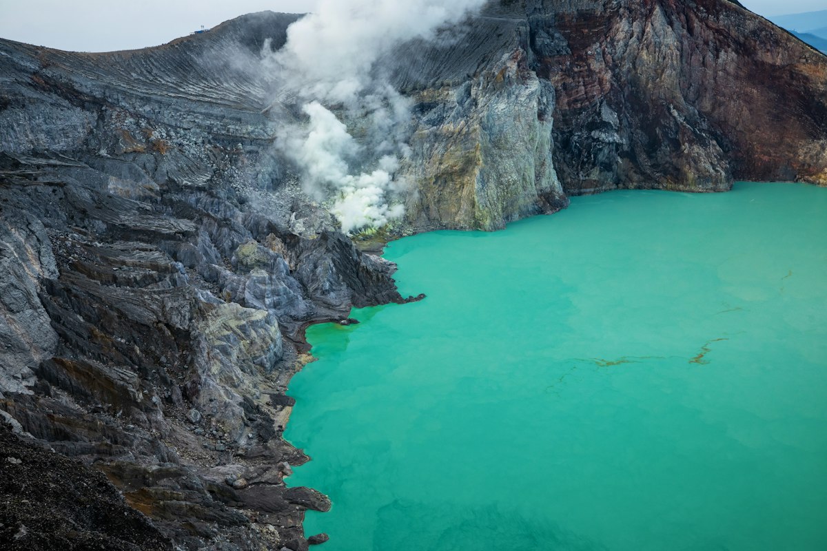 Turquoise acidic crater lake ringed by steep rock walls with sulfur steam drifting across it
