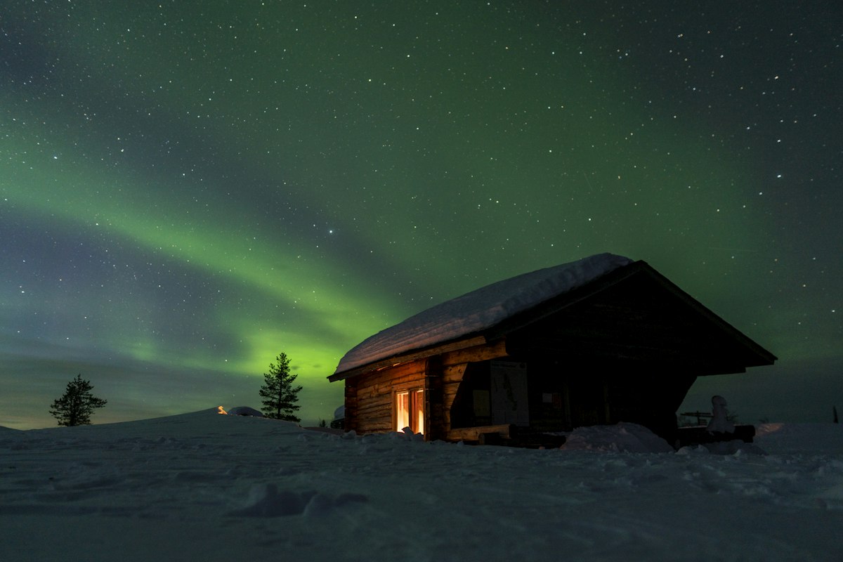 Wooden cabin standing in deep snow under a green aurora filling the night sky