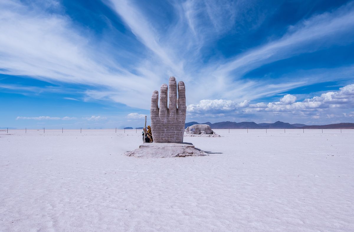 Giant concrete hand sculpture rising out of flat pale desert ground under a wide blue sky