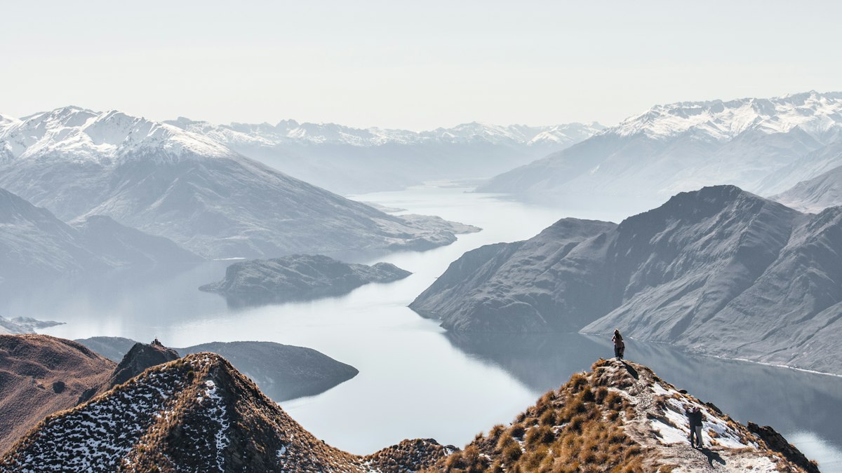 A lone hiker standing on a narrow ridge above a lake with mist and snow covered peaks in the distance