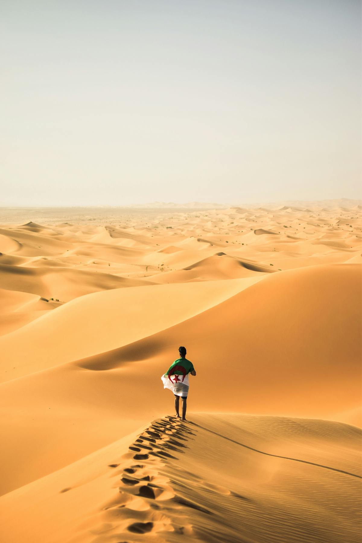 A person walking alone across vast golden sand dunes of the Sahara Desert with footprints trailing behind under hazy skies