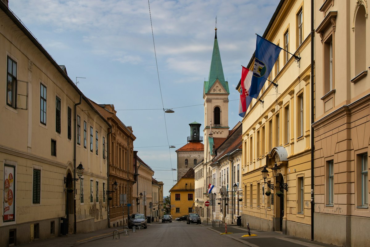 Quiet street in Zagreb's Upper Town lined with pastel buildings and flags, with a church tower at the end