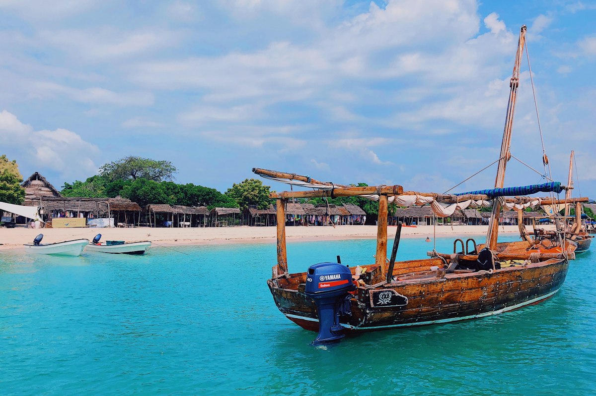 Traditional wooden dhow boat moored in shallow turquoise water in front of a Zanzibar beach with thatched roof huts